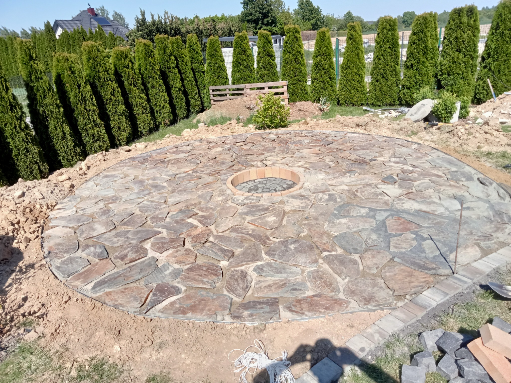 Okrągła powierzchnia wyłożona nieregularnymi kamieniami z centralnym paleniskiem otoczonym cegłą, w trakcie wykańczania obrzeża z kostki brukowej, na tle żywopłotu z tuj.
