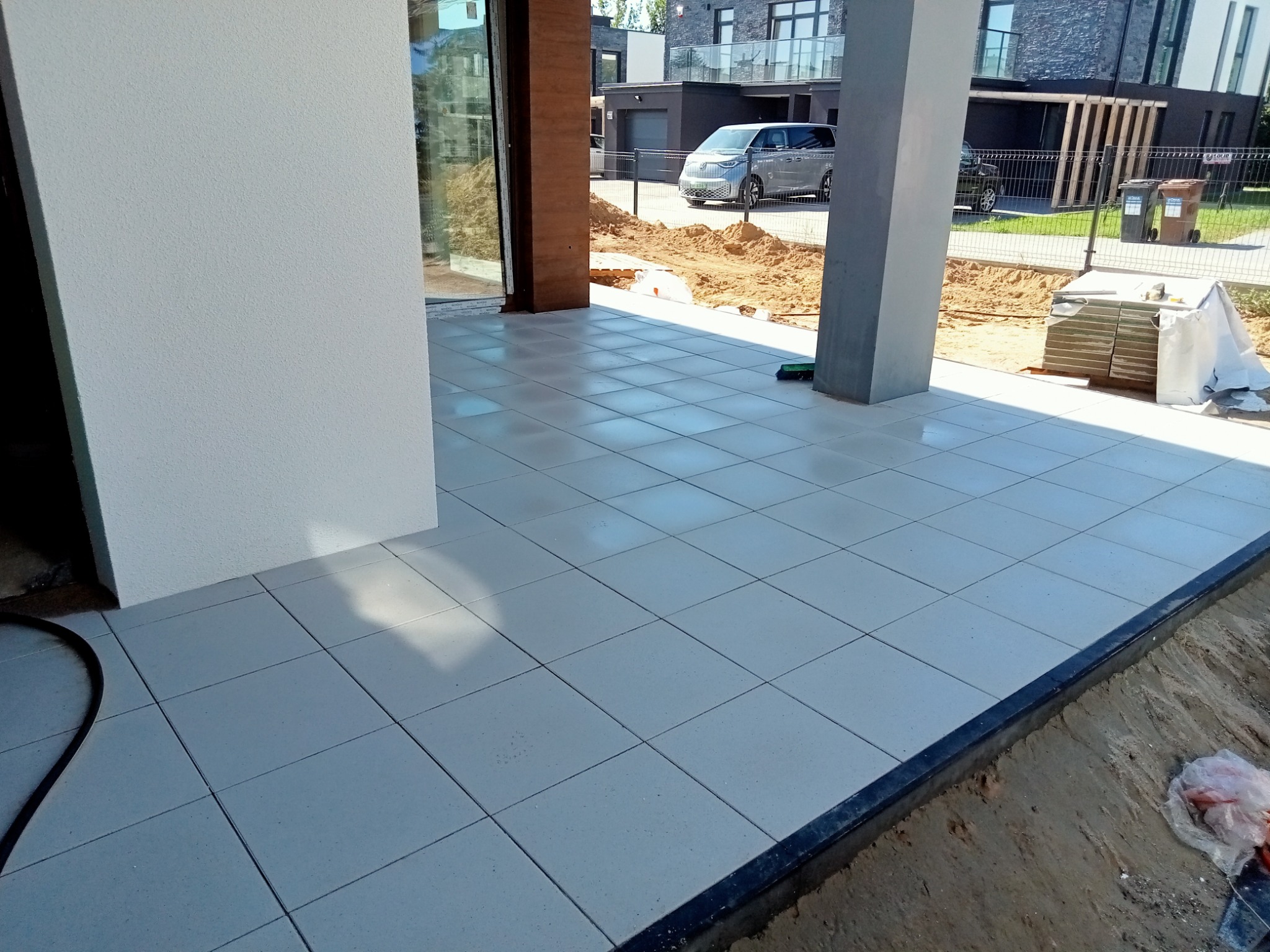 Jasne, kwadratowe płytki ułożone na tarasie domu jednorodzinnego, widoczne przygotowania do dalszych prac ziemnych w tle.