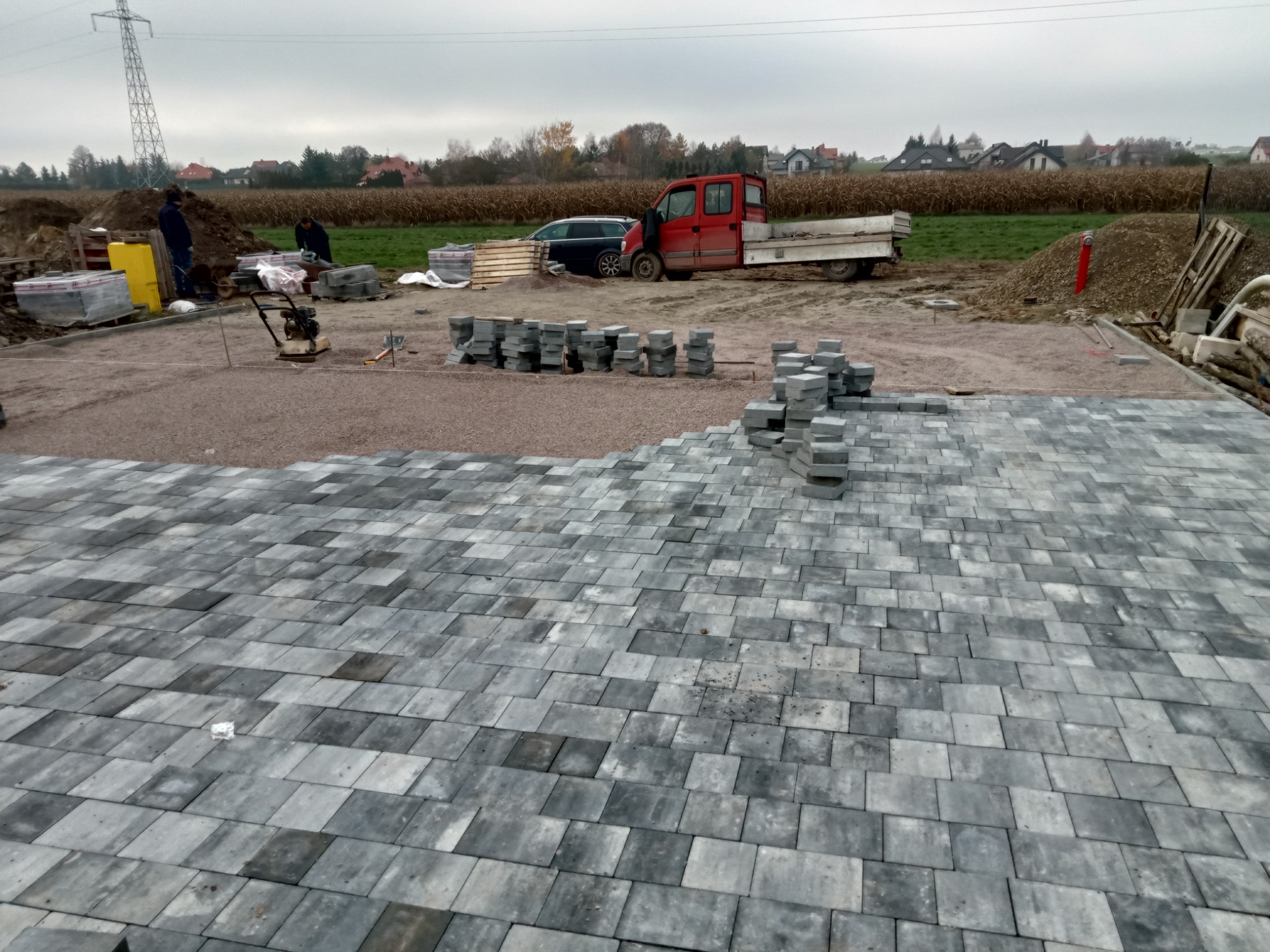 Trwa układanie szarej kostki brukowej w Lublinie. Widoczny fragment ułożonej nawierzchni, przygotowane podłoże z gruzem, stosy kostki, pracownicy oraz czerwony samochód dostawczy w tle.