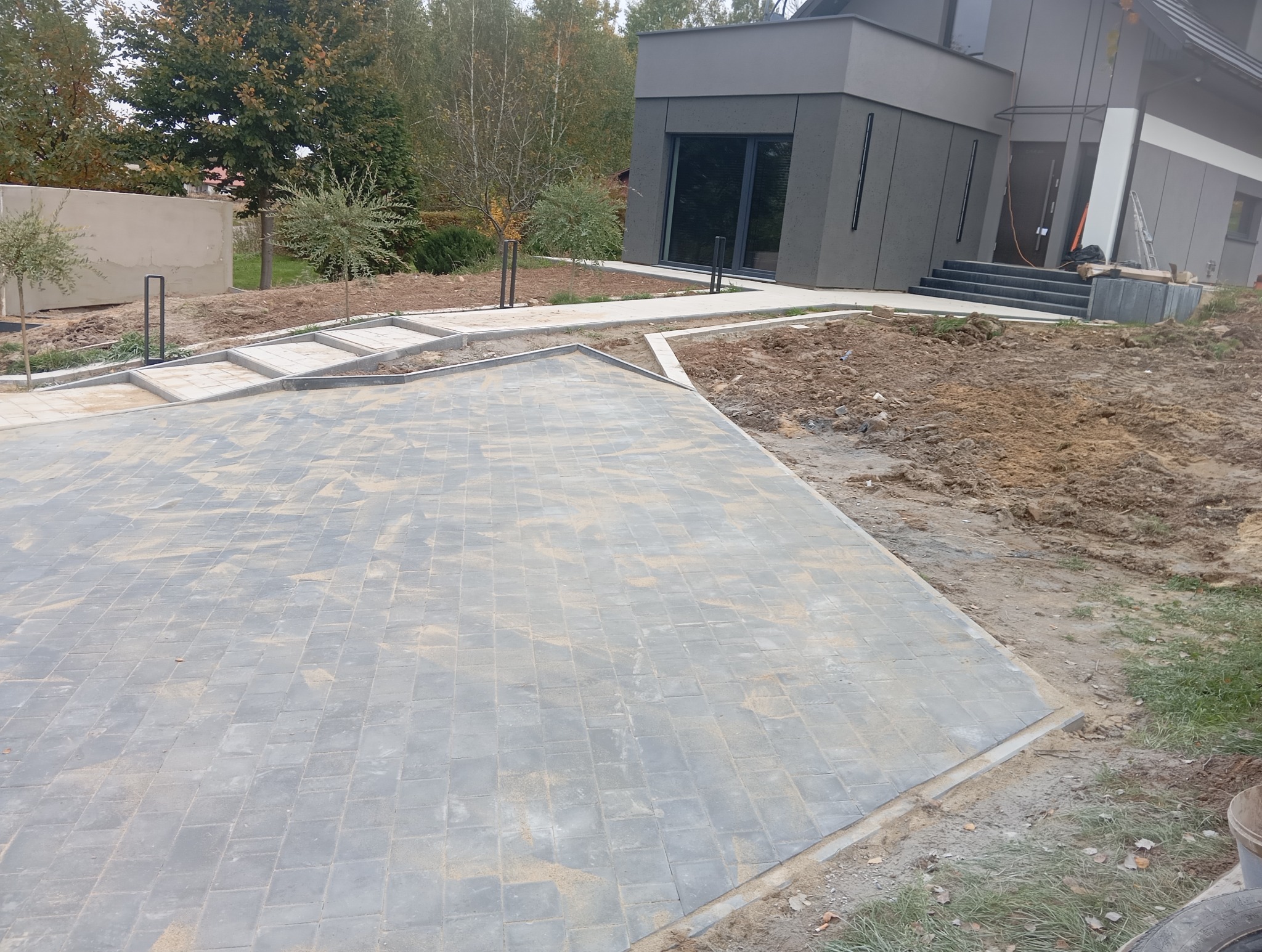 Nowoczesna posesja z szarej kostki brukowej ułożonej w geometryczny wzór. Widoczne obrzeża i fragment domu w tle. Świeżo ułożona nawierzchnia.