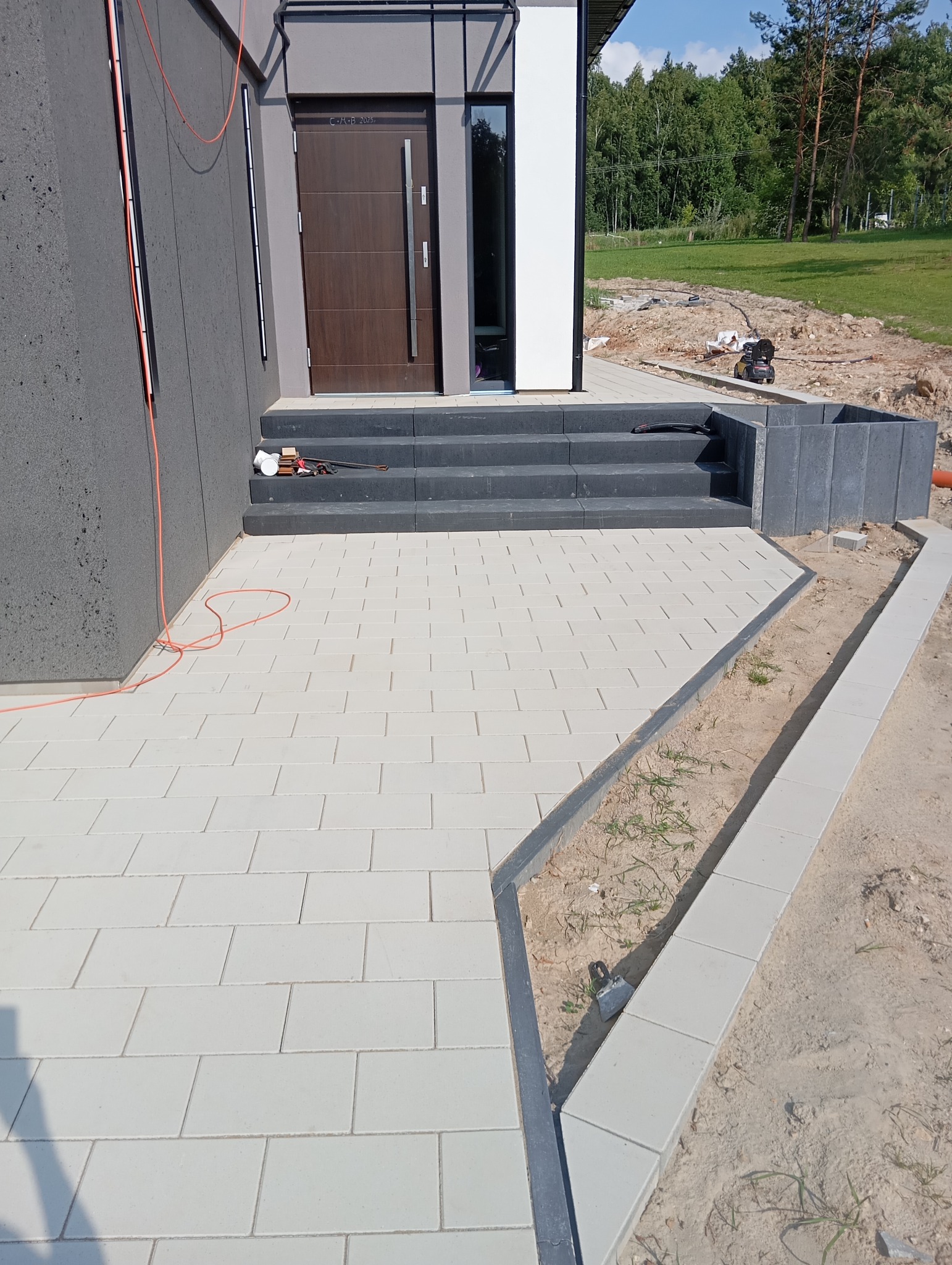 Jasnoszara kostka brukowa ułożona przed nowoczesnym domem z ciemnymi schodami wejściowymi. Widoczne obrzeża i fragment trawnika w tle.