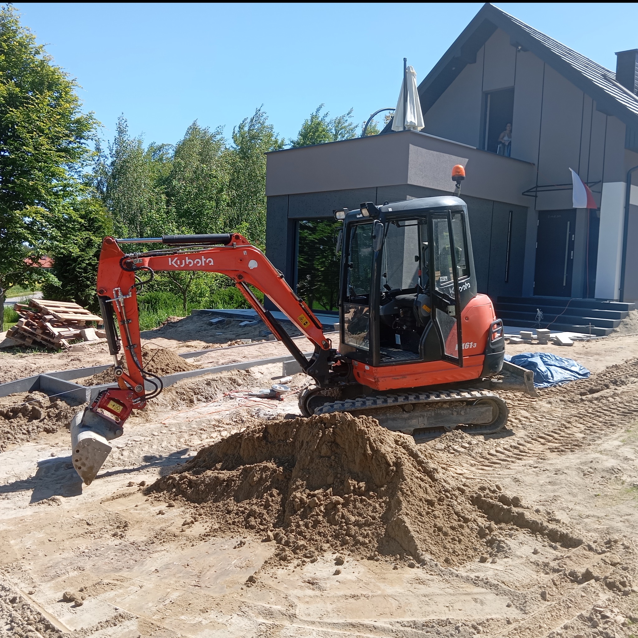 Pomarańczowa minikoparka Kubota na placu budowy, przygotowuje teren pod kostkę brukową. W tle nowoczesny dom z osobą w oknie i palety z materiałami.