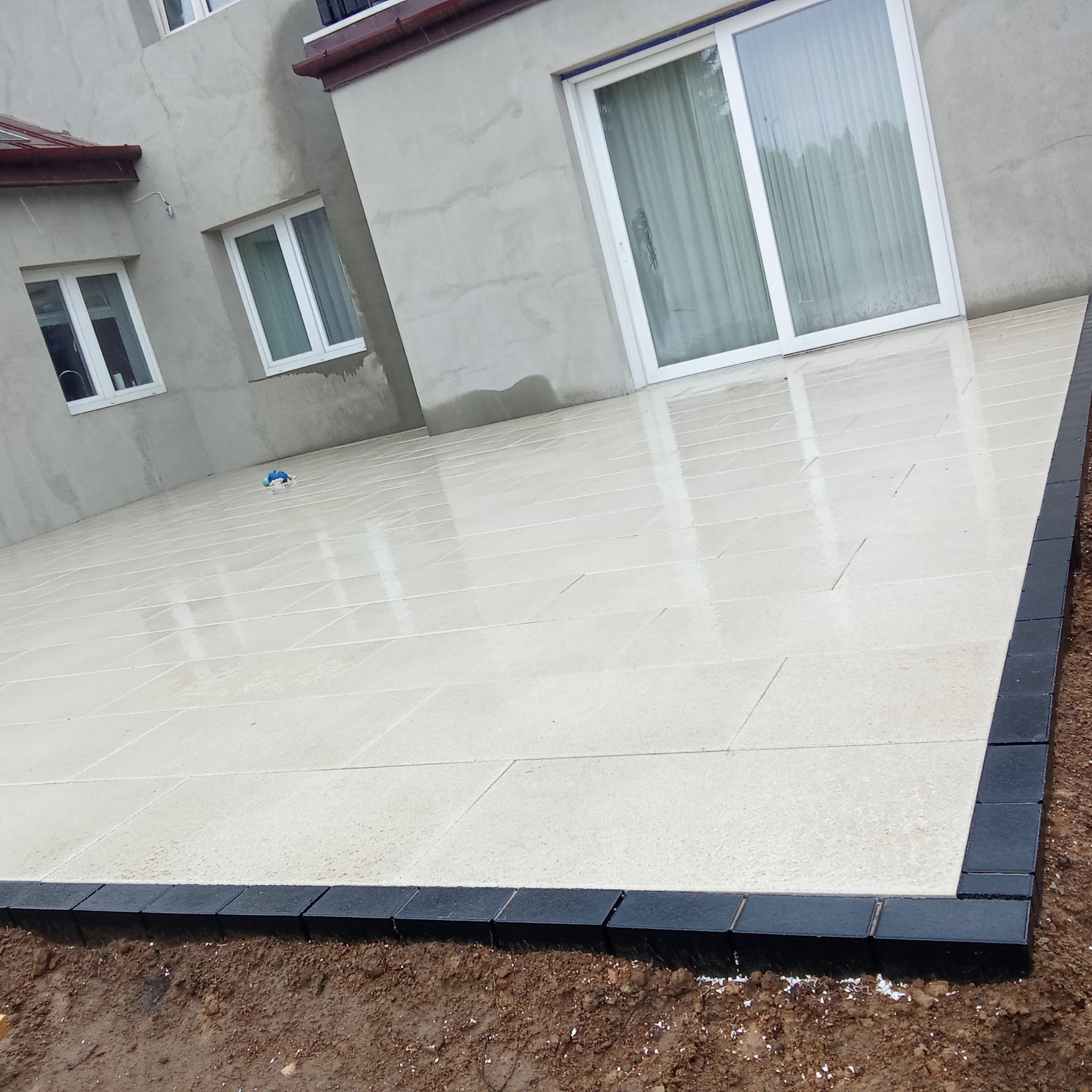 Jasna, prostokątna kostka na tarasie domu z czarnym obramowaniem. Widoczne okna i drzwi balkonowe. Kostka lekko mokra, odbija światło.