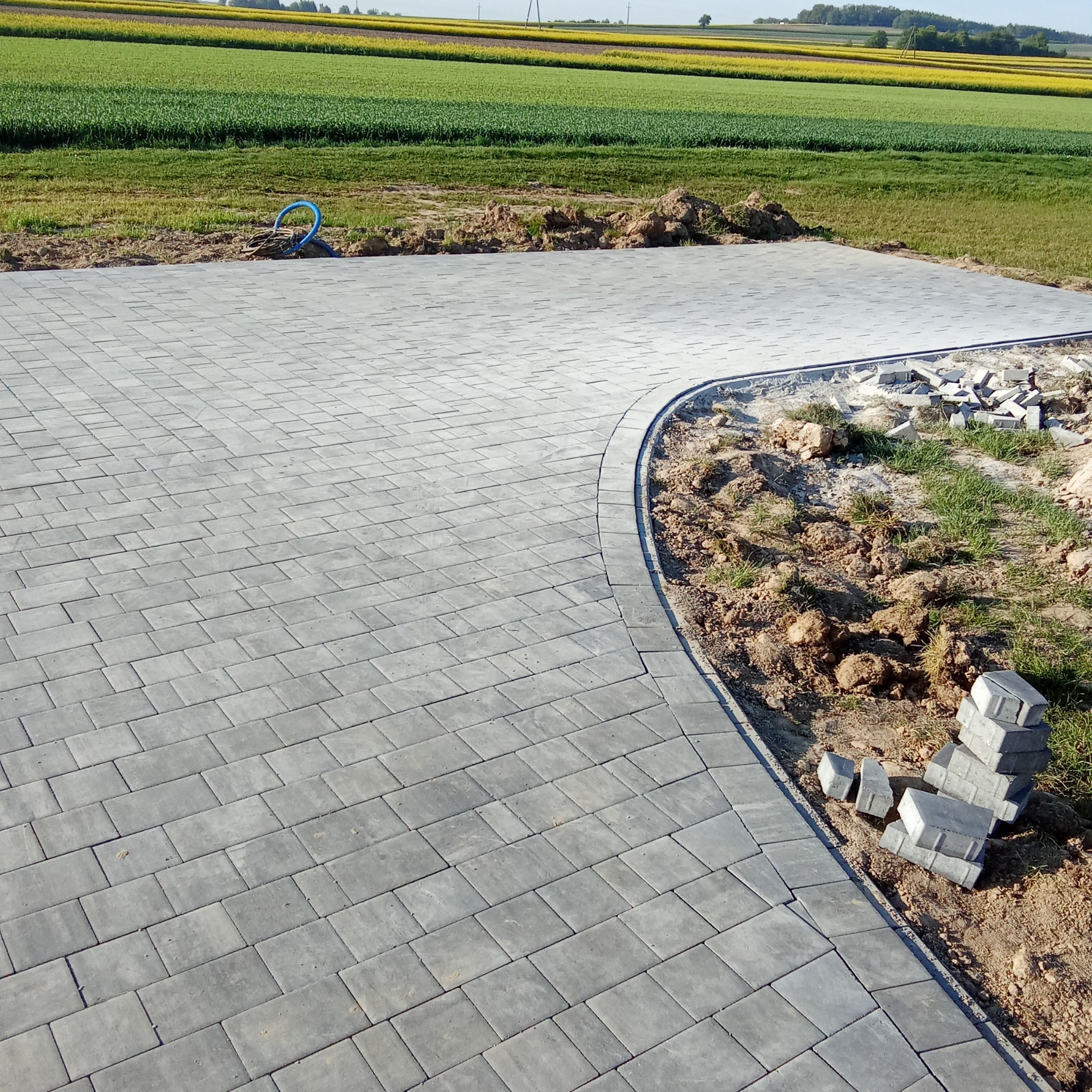 Ułożona szara kostka brukowa na podjeździe, z widocznym fragmentem niezabudowanym i stosem kostki. W tle pole uprawne i błękitne niebo.