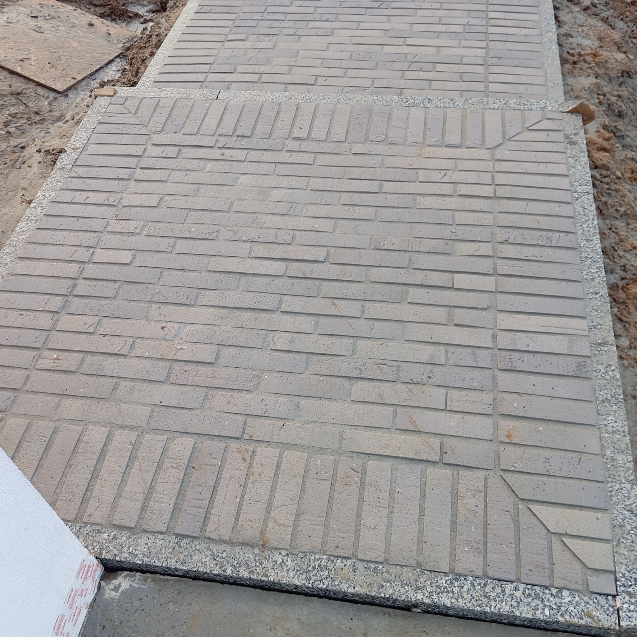 Szara kostka brukowa ułożona w geometryczny wzór, obramowana drobnym grysem. Widoczne fragmenty podłoża i materiałów budowlanych. Ujęcie z góry.