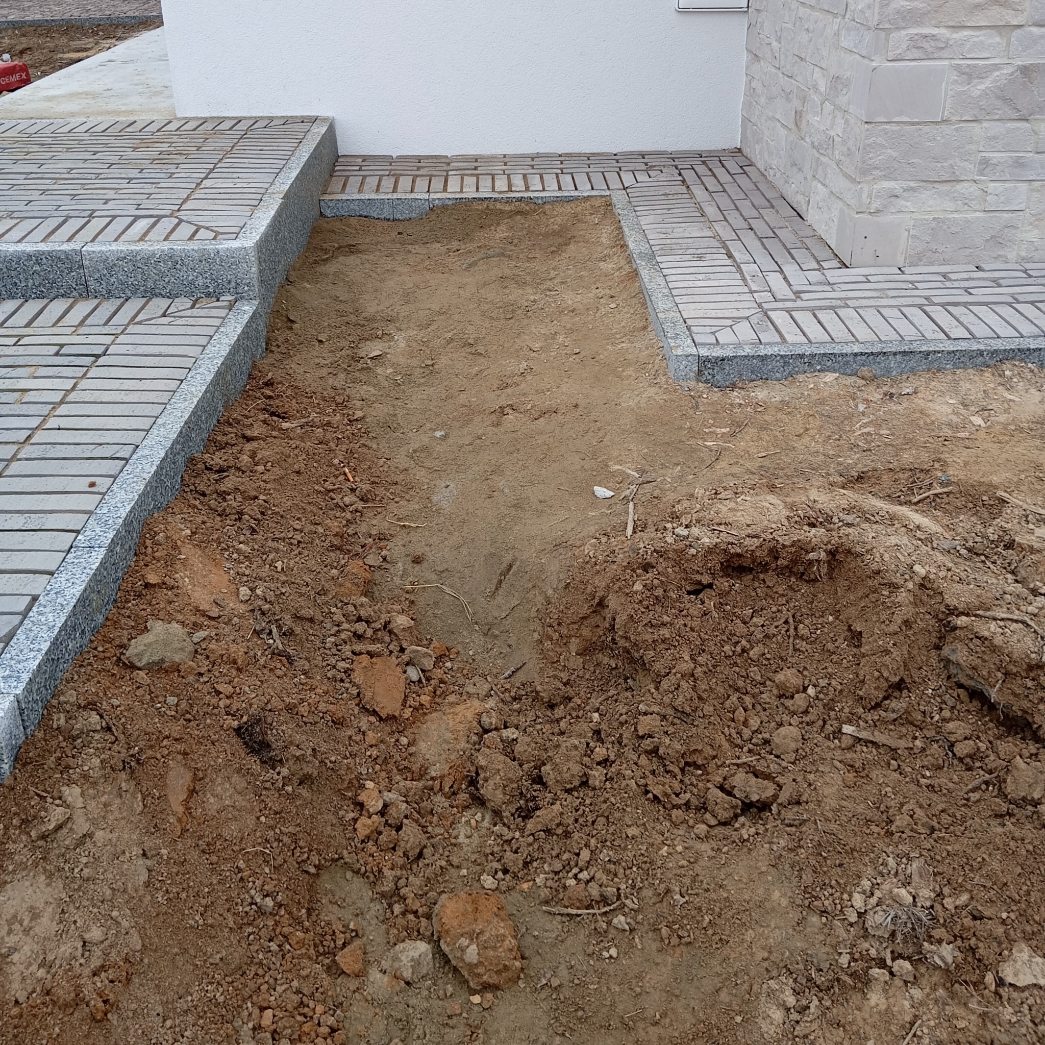 Fragment podjazdu z kostki granitowej w trakcie budowy. Częściowo ułożona kostka, obok wykopana ziemia. Widoczne obrzeża i fragment elewacji budynku.