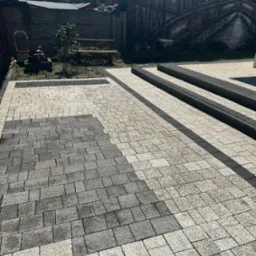 Układanie dwukolorowej kostki brukowej na posesji, widoczny fragment nieułożonej kostki, w tle budynki i zieleń.