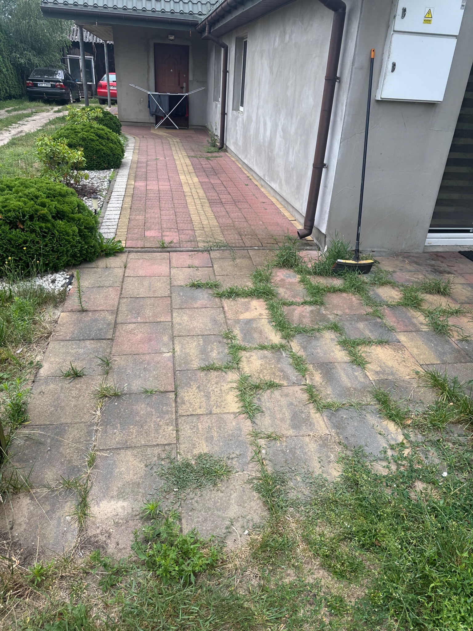 Zarośnięta kostka brukowa przed domem z widoczną miotłą opartą o ścianę, sugerująca prace porządkowe w toku.