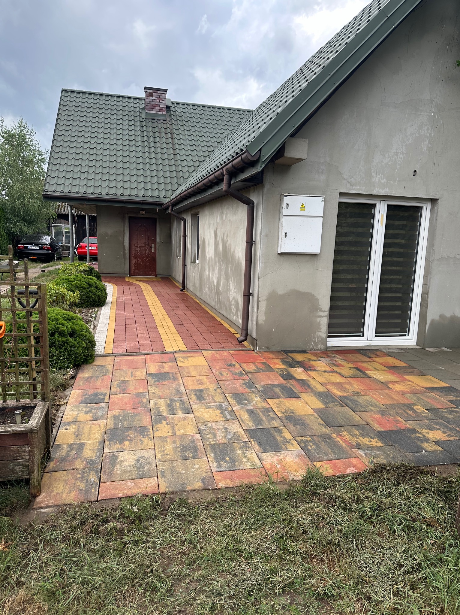 Kolorowa kostka brukowa prowadząca do wejścia domu z zielonym dachem, widoczne przyłącze elektryczne na elewacji i fragment ogrodu