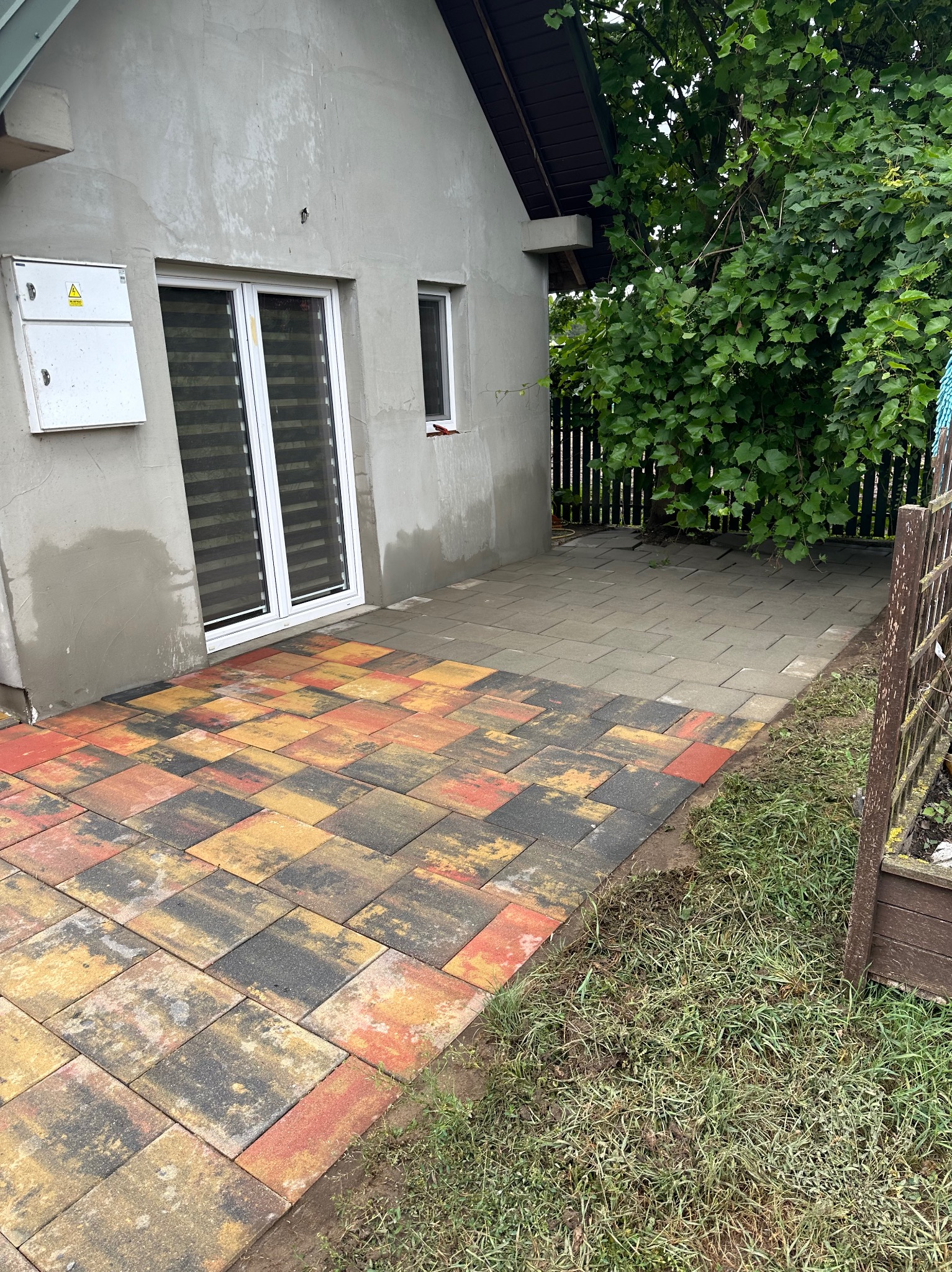Ułożona, wielobarwna kostka brukowa (czerwona, żółta, czarna) przy ścianie domu z białymi oknami i zielonymi krzewami w tle. Część kostki jest szara, a część kolorowa.