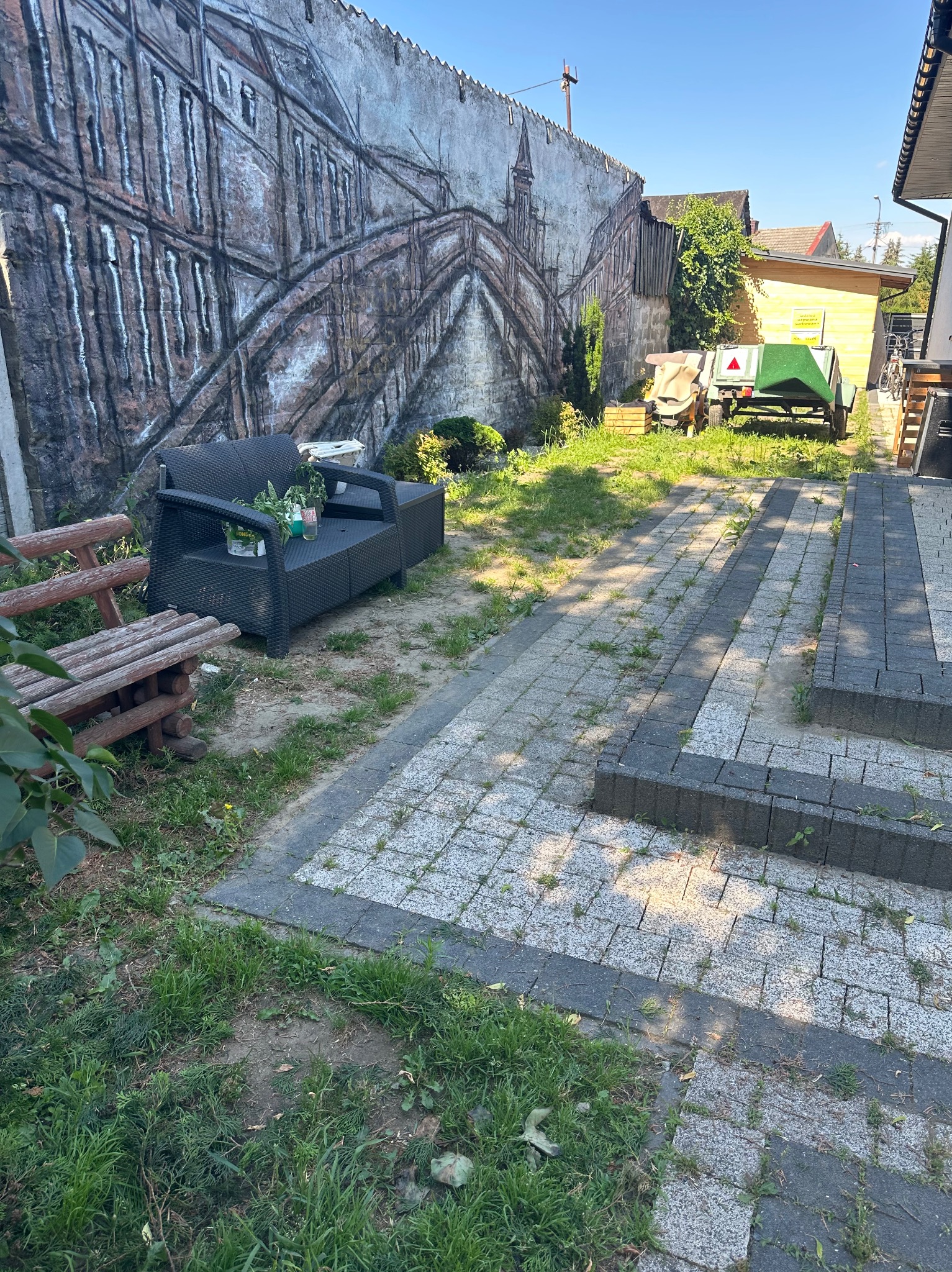 Szara kostka brukowa z trawą wyrastającą między szczelinami, obok drewniana ławka i czarny rattanowy komplet wypoczynkowy z roślinami, na betonowym murze namalowany most.