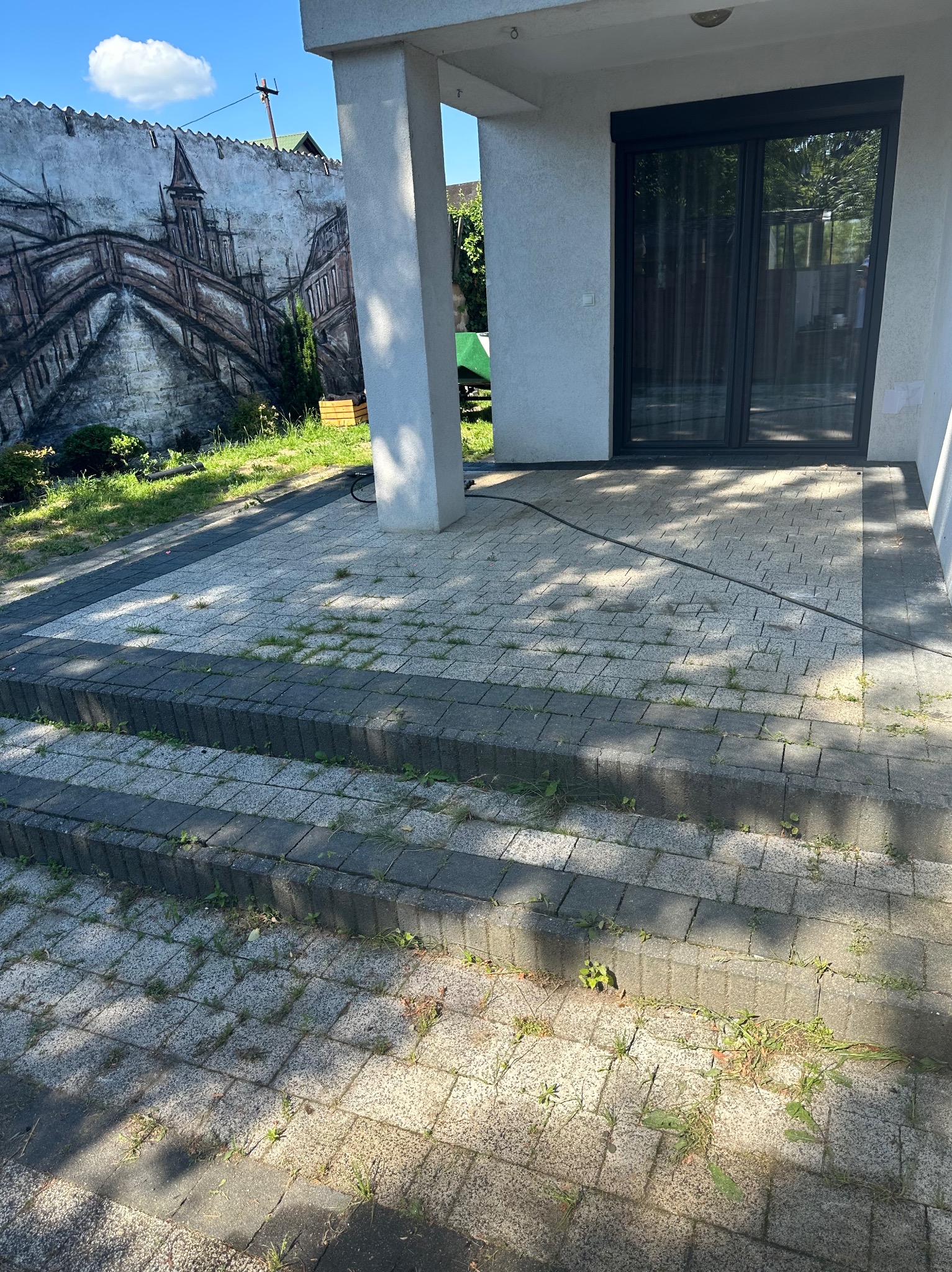 Schody i kostka brukowa na posesji z widocznymi chwastami, na ścianie graffiti z motywem architektonicznym.