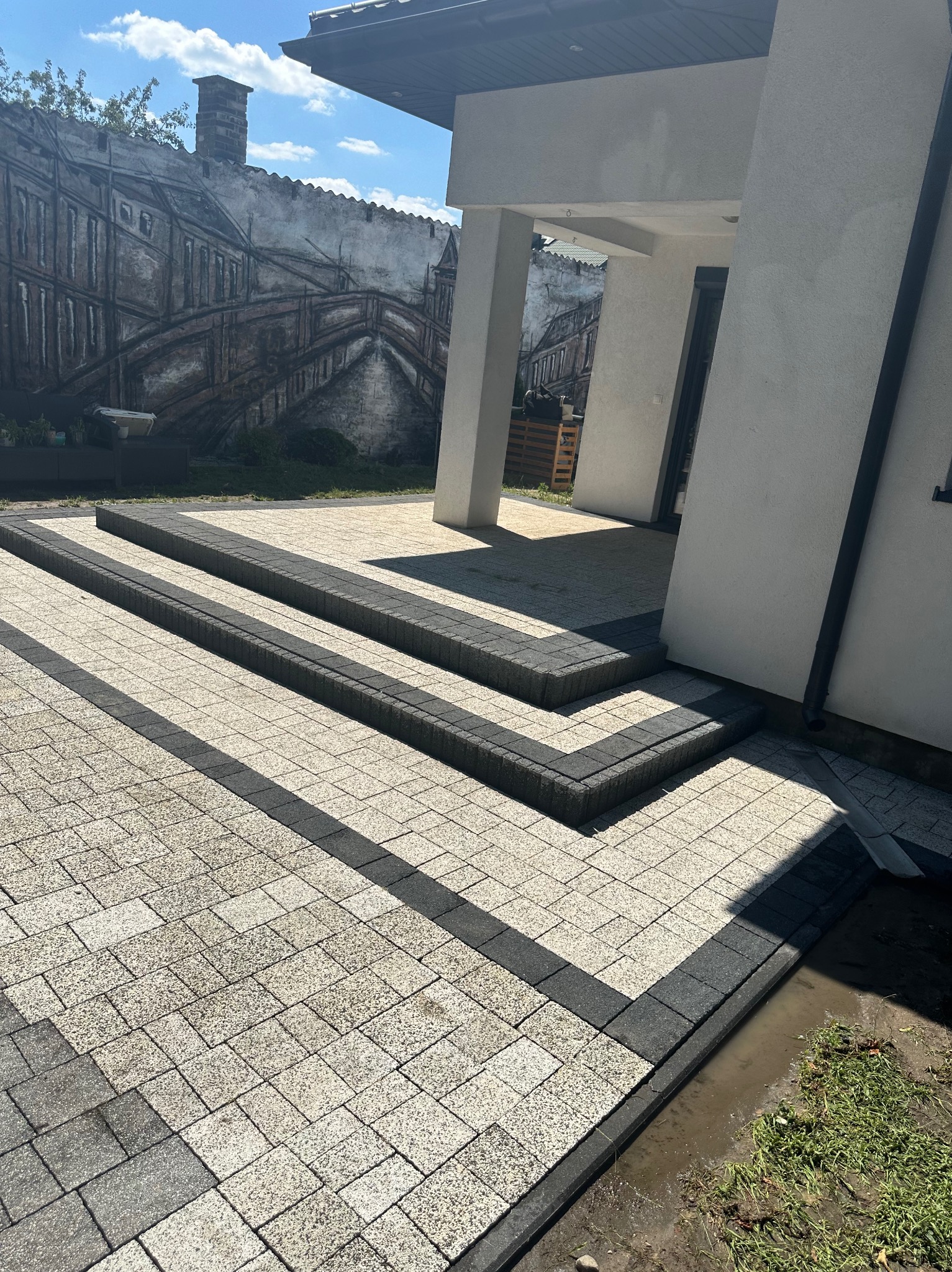 Ułożona kostka brukowa w odcieniach szarości i beżu tworząca schody wejściowe do nowoczesnego domu z muralem na murze w tle.