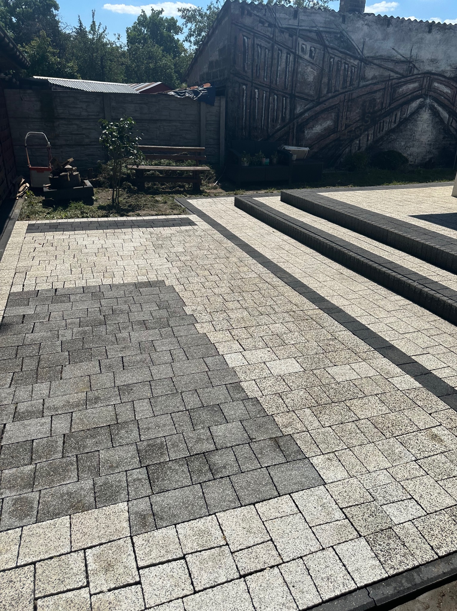 Układanie dwukolorowej kostki brukowej na posesji, widoczny fragment nieułożonej kostki, w tle budynki i zieleń.