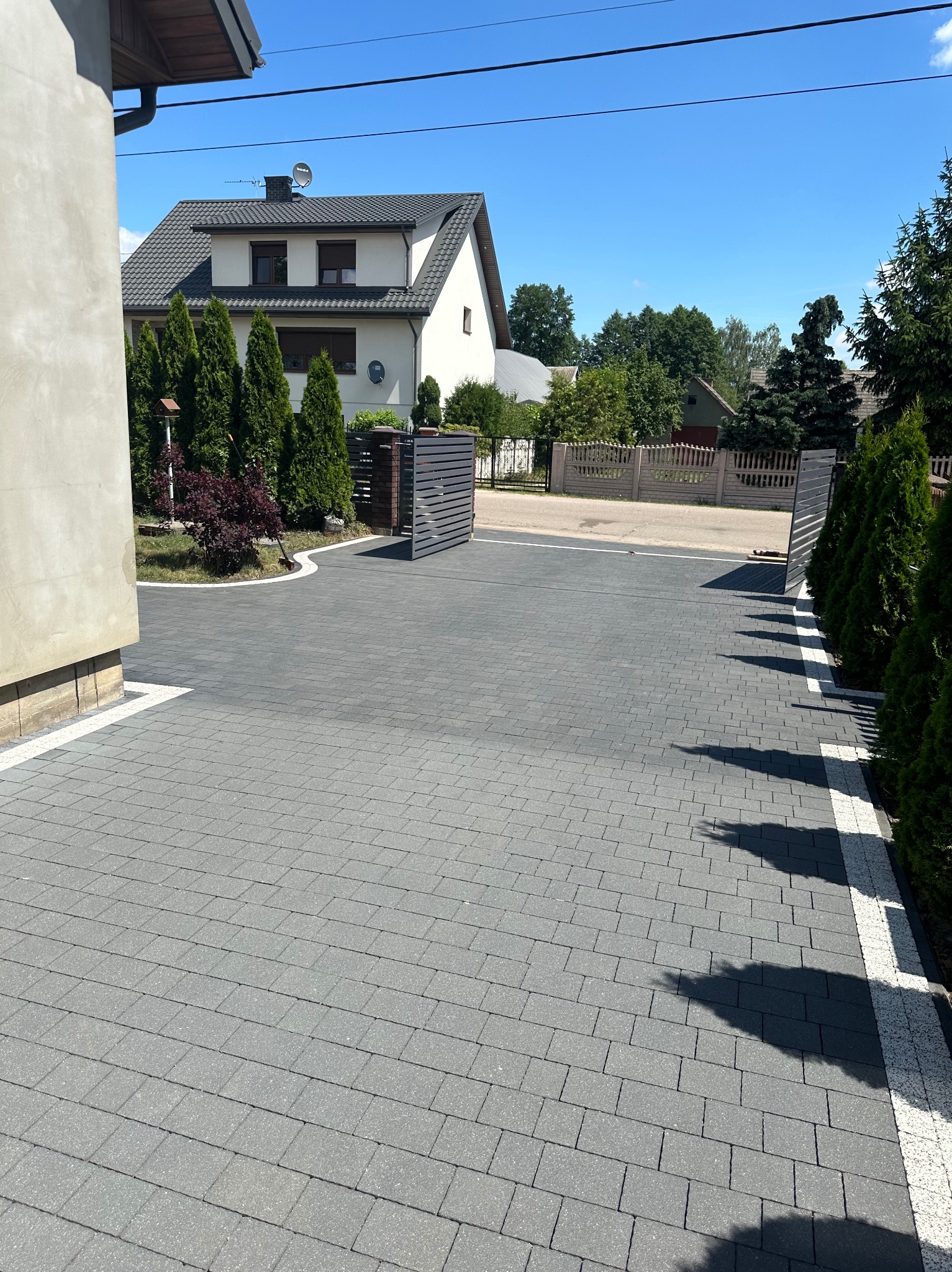 Nowoczesny podjazd z szarej kostki brukowej ułożonej we wzór cegły, obrzeża z białego kamienia, tuje i brama w tle, dom jednorodzinny z szarą dachówką w słoneczny dzień.
