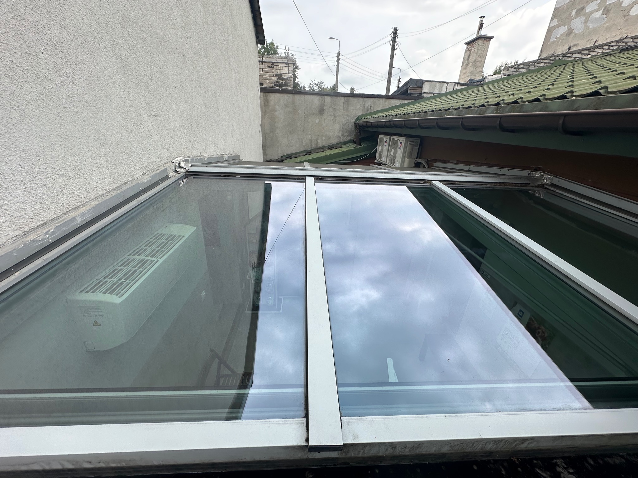 Szklany dach z widocznymi zabrudzeniami, odbijający pochmurne niebo, z fragmentem elewacji i dachu sąsiedniego budynku w tle.