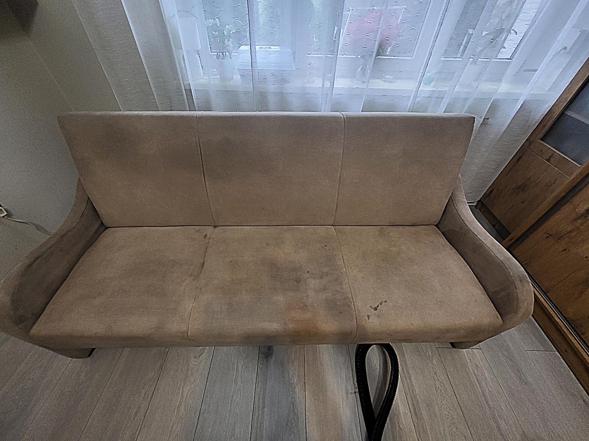 Beżowa kanapa z widocznymi plamami na siedzisku, częściowo przysłonięta wężem odkurzacza piorącego, na tle okna z firanką i drewnianego elementu wystroju.