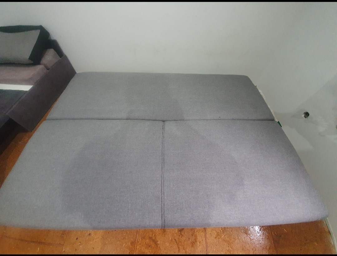 Szary narożnik z widocznymi szwami na siedzisku, ustawiony na drewnianej podłodze, z fragmentem innego mebla w tle.