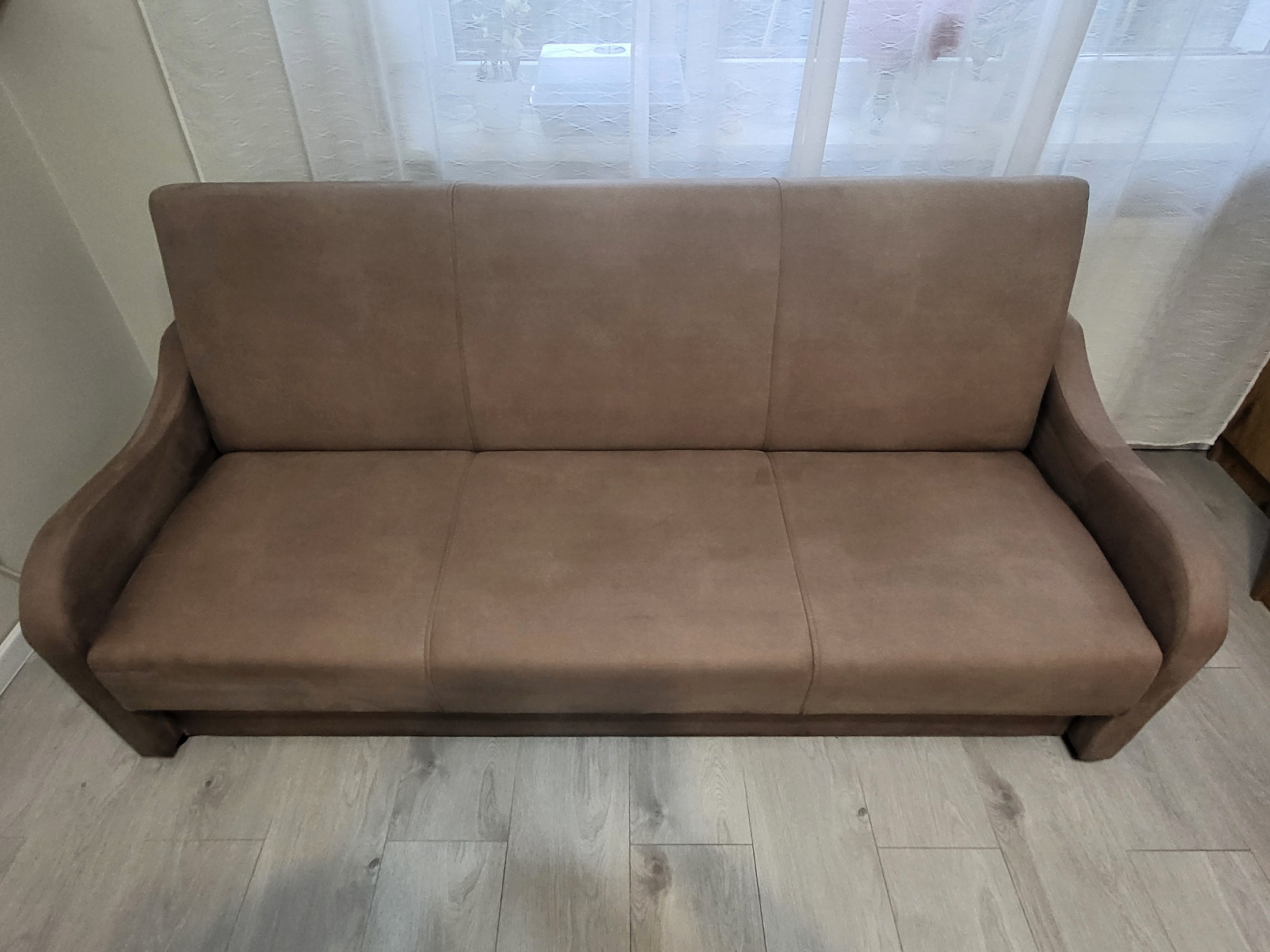 Brązowa, rozkładana kanapa z trzema siedziskami, stojąca na panelach podłogowych, oświetlona naturalnym światłem z okna zza firanki.