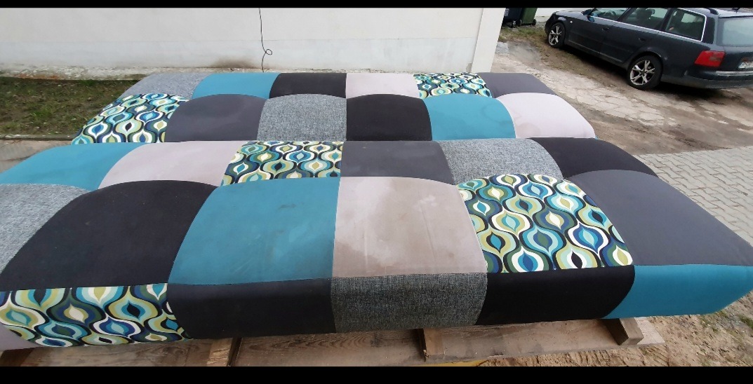 Wielobarwna, patchworkowa sofa, z widocznymi plamami na jasnym panelu, stojąca na zewnątrz przy szarym samochodzie.