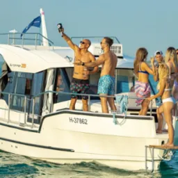 Grupa młodych osób w strojach kąpielowych świętuje na pokładzie białej motorówki na tle błękitnej wody i oddalonego brzegu; jeden mężczyzna wznosi toast butelką, inny trzyma kieliszek szampana.