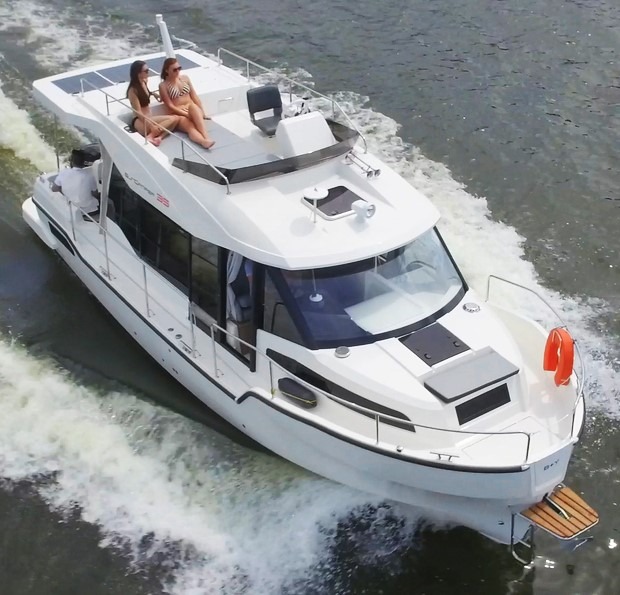 Biały jacht motorowy SunCamper 35 Flybridge płynie po wodzie, z dwiema osobami opalającymi się na górnym pokładzie i jedną osobą siedzącą na niższym pokładzie.