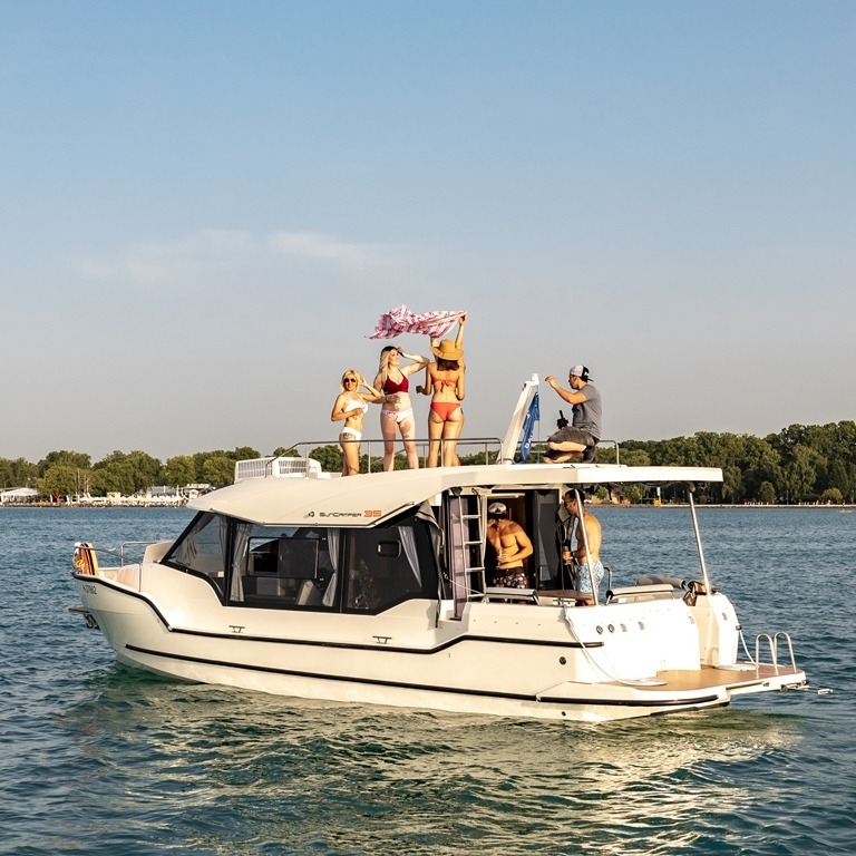 Grupa osób w strojach kąpielowych na pokładzie białego jachtu motorowego Sun Camper 35 na tle spokojnej wody i zieleni drzew na brzegu.