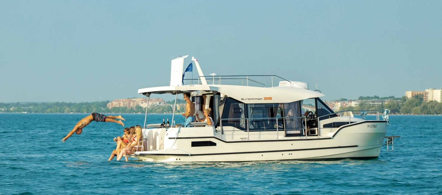 Grupa osób relaksuje się na pokładzie białego jachtu SunCamper 35; jeden mężczyzna skacze do wody, w tle zabudowania miejskie.