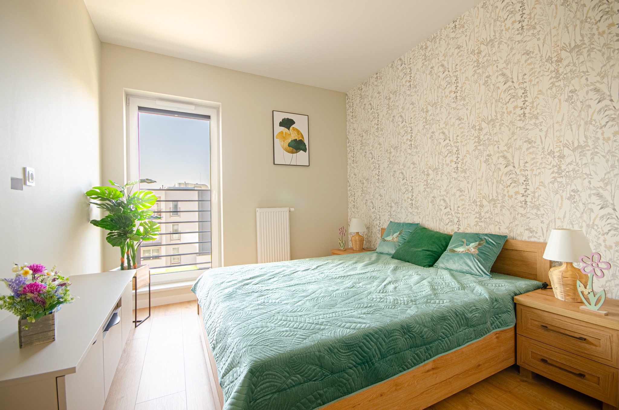 Jasna sypialnia z łóżkiem o drewnianej ramie, przykrytym zieloną narzutą z pikowanym wzorem liści, wzorzysta tapeta w odcieniach beżu, kwiaty w doniczce na szafce nocnej i para roślin doniczkowych...