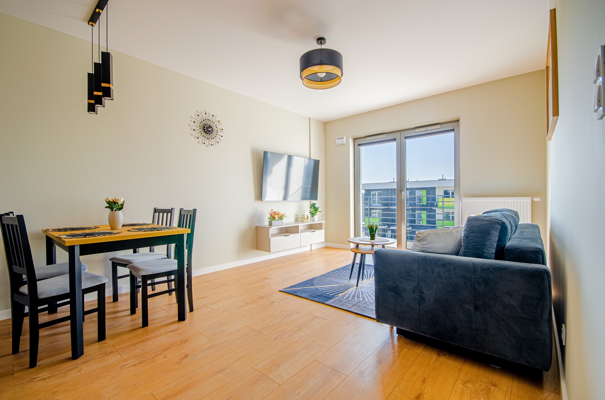 Jasne wnętrze salonu z drewnianą podłogą, szarą sofą, stołem z czarnymi krzesłami, telewizorem na ścianie i dużym oknem balkonowym z widokiem na budynek.