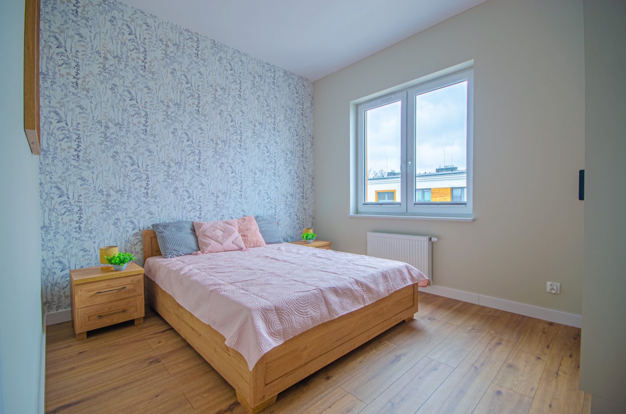 Jasne wnętrze sypialni z drewnianym łóżkiem, pastelową narzutą i wzorzystą tapetą na jednej ścianie. Widoczne dwie szafki nocne z lampkami i kwiatami, drewniana podłoga oraz okno z widokiem...