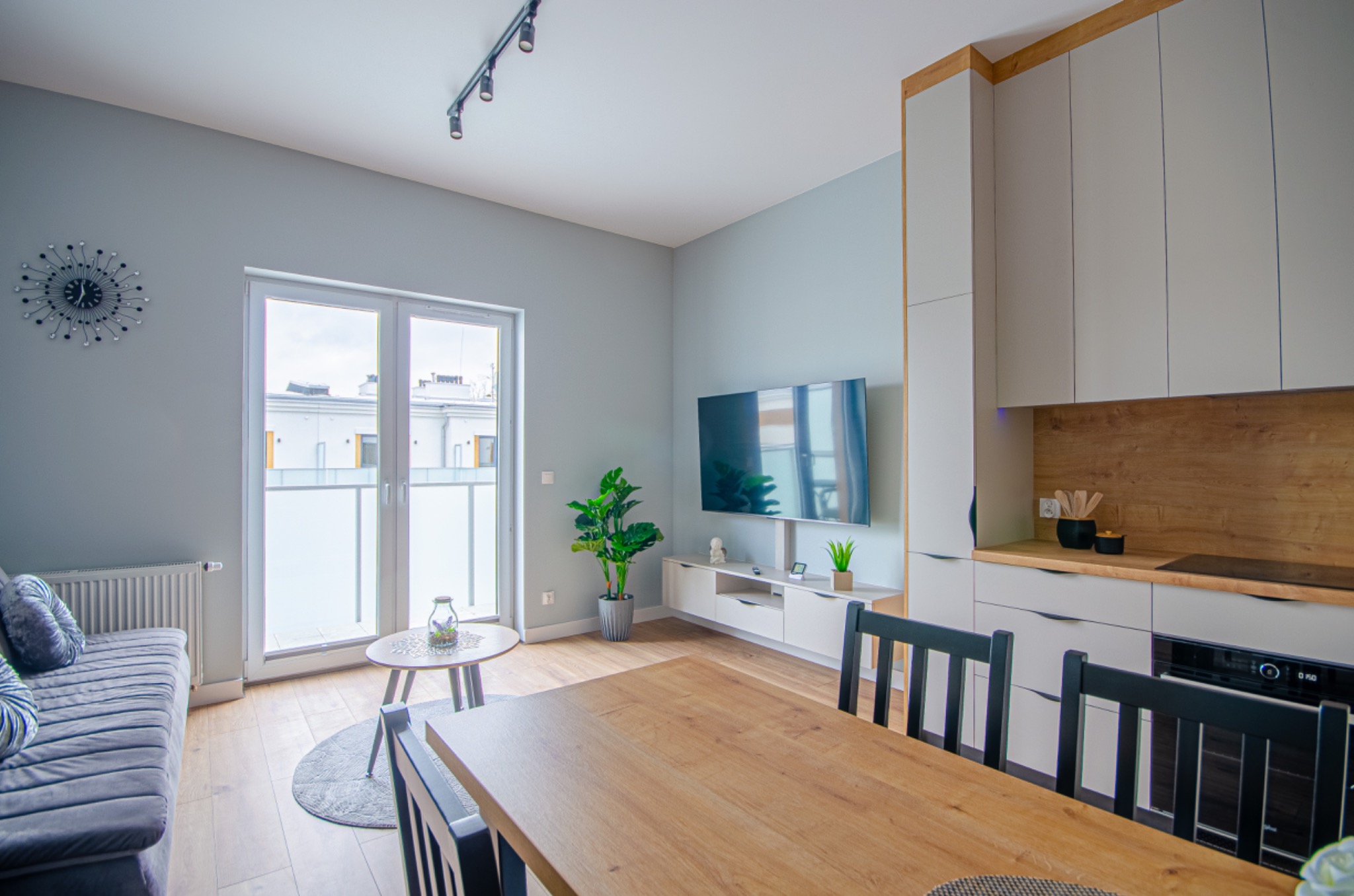 Jasne, nowoczesne wnętrze mieszkania z aneksem kuchennym: drewniany stół na pierwszym planie, szara sofa z poduszkami, telewizor na ścianie, zabudowa kuchenna z drewnianym blatem i sprzętem AGD.