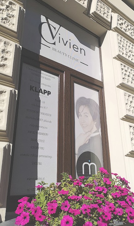 Elegancka witryna salonu urody Vivien Beauty Clinic z informacjami o zabiegach Klapp Cosmetics, makijażu permanentnym, stylizacji brwi i szkoleniach, ozdobiona skrzynką z kwitnącymi różowymi...