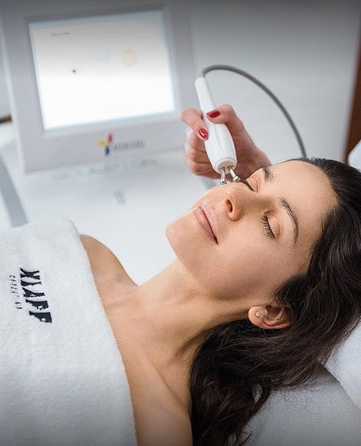 Kobieta z zamkniętymi oczami podczas zabiegu na twarz z użyciem urządzenia z elektrodami, na białym tle widoczny ekran urządzenia.