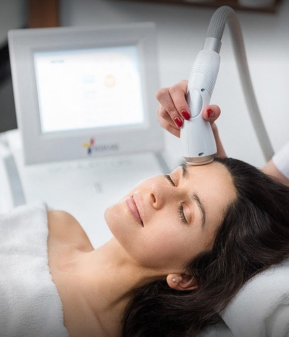 Kobieta leżąca na leżance poddawana zabiegowi na twarz przy użyciu urządzenia z głowicą, widoczny ekran urządzenia w tle.