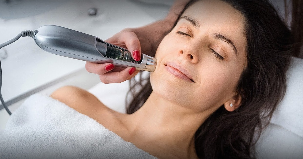 Zabieg na twarz kobiety wykonywany srebrnym urządzeniem z przezroczystym elementem, czerwony manicure, biały ręcznik, delikatny makijaż.