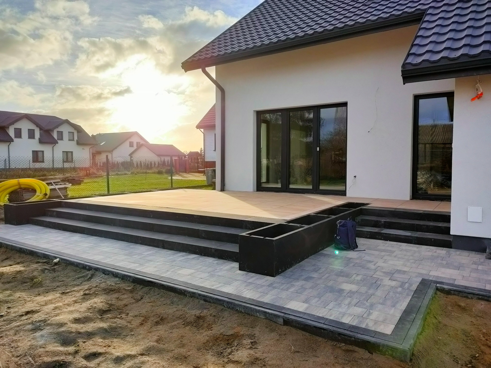 Wykończony taras z kostki brukowej w odcieniach szarości, z czarnymi schodami i donicami, przylegający do nowoczesnego domu z białymi ścianami i ciemną dachówką, w tle zabudowa jednorodzinna...