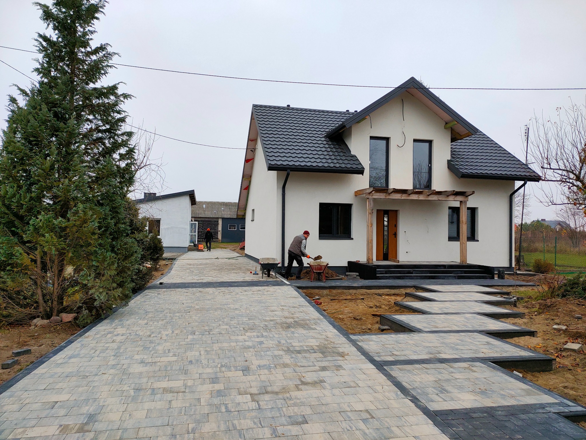 Układanie kostki brukowej na podjeździe do nowoczesnego domu z czarnym dachem, widoczny brukarz z taczką i narzędziami w trakcie pracy, częściowo ułożona nawierzchnia z szaro-czarnej kostki.