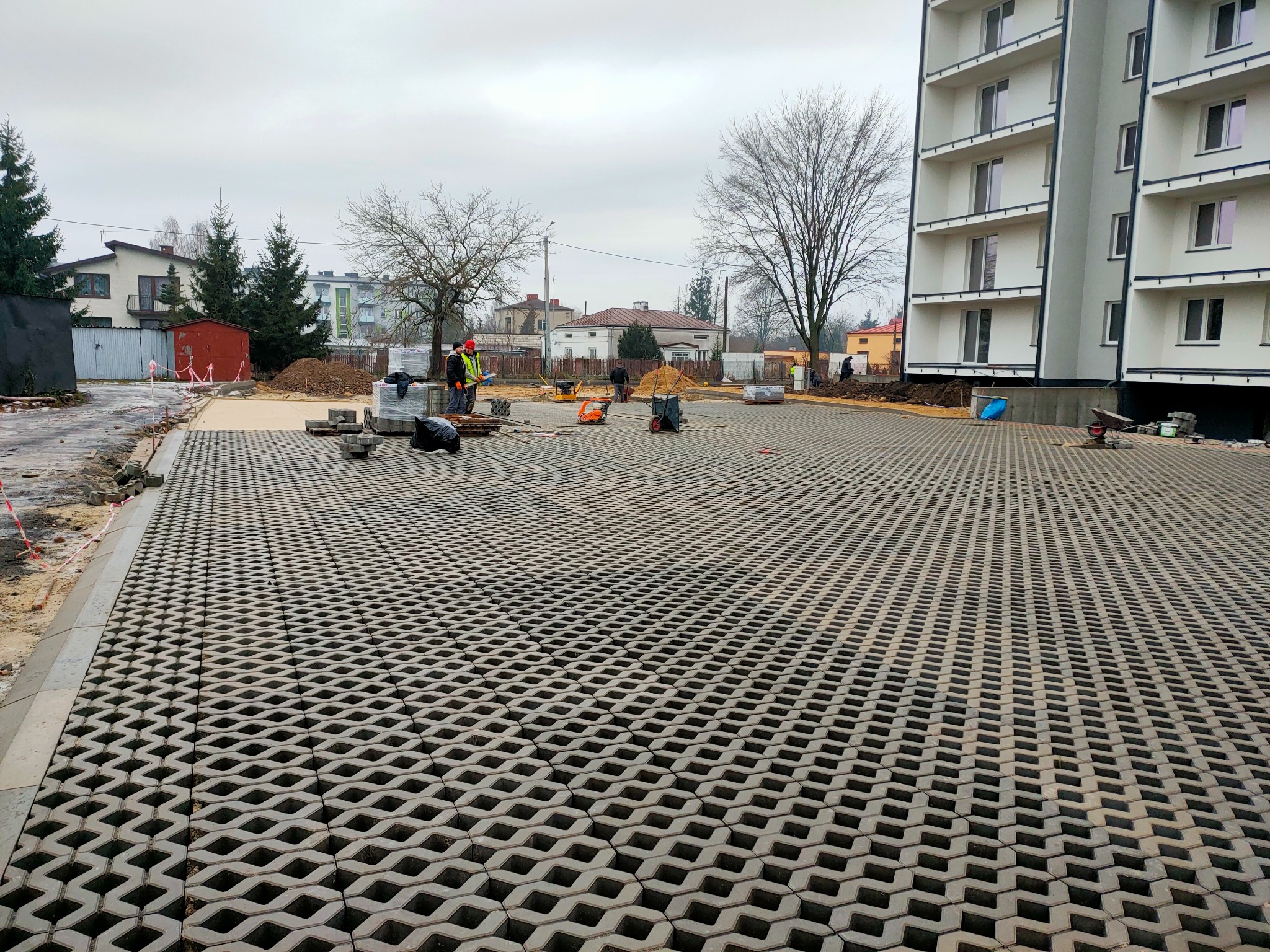 Ułożona, ażurowa nawierzchnia z betonowych płyt na tle budynków mieszkalnych i pracujących robotników, widoczne materiały budowlane i narzędzia.
