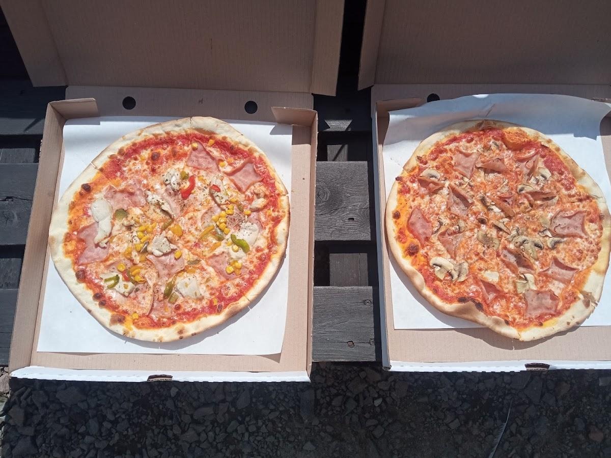 Dwie pizze w kartonowych pudełkach na drewnianym stole: jedna z szynką, kurczakiem, kukurydzą i papryką, druga z szynką i pieczarkami.