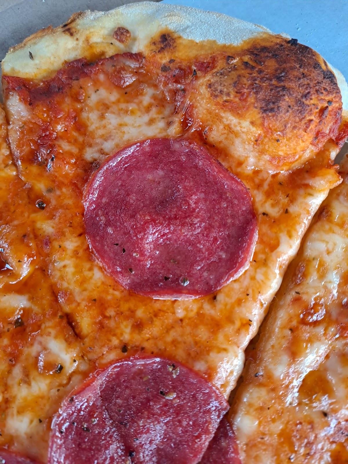 Fragment pizzy z salami, widoczne błyszczące krople tłuszczu na salami i serze, lekko przypieczone ciasto.