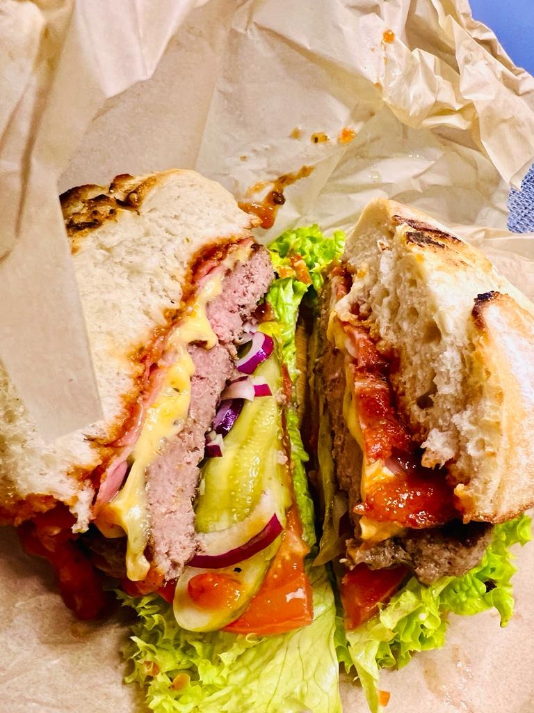 Przekrojony burger z wołowiną, serem cheddar, piklami, czerwoną cebulą, pomidorem i sosem, podawany w papierowej torbie.