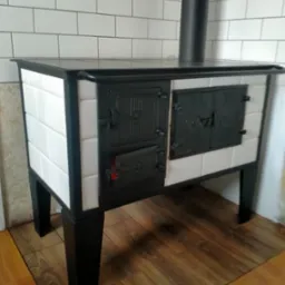 Kuchenny piec kaflowy z czarnymi drzwiczkami i rurą odprowadzającą dym, obudowany białymi kaflami, ustawiony na drewnianej podłodze.