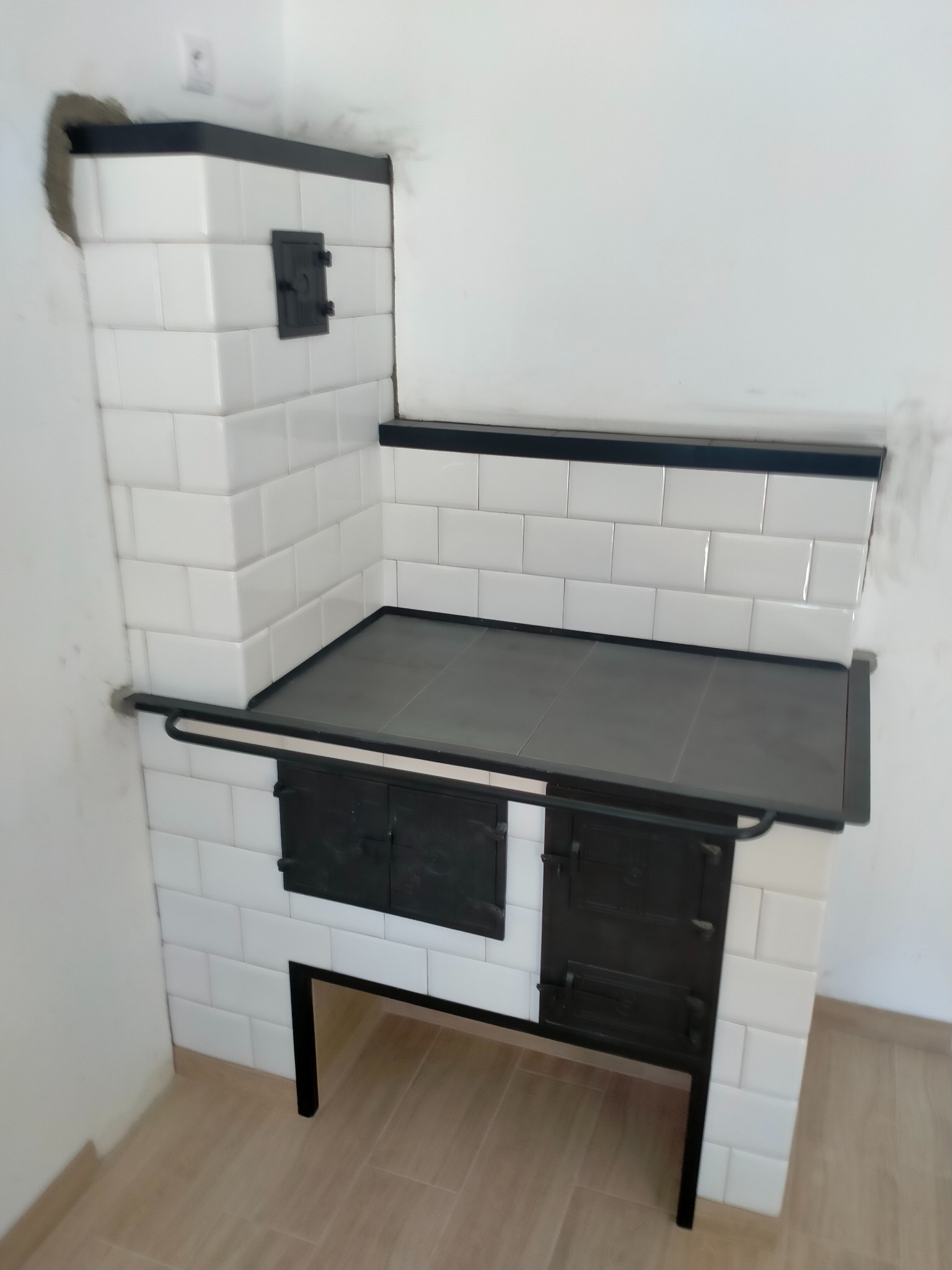 Biały piec kaflowy z czarnymi drzwiczkami i blatem, wykończony czarnymi listwami, w trakcie montażu, widoczne niedociągnięcia w tynkowaniu ściany.