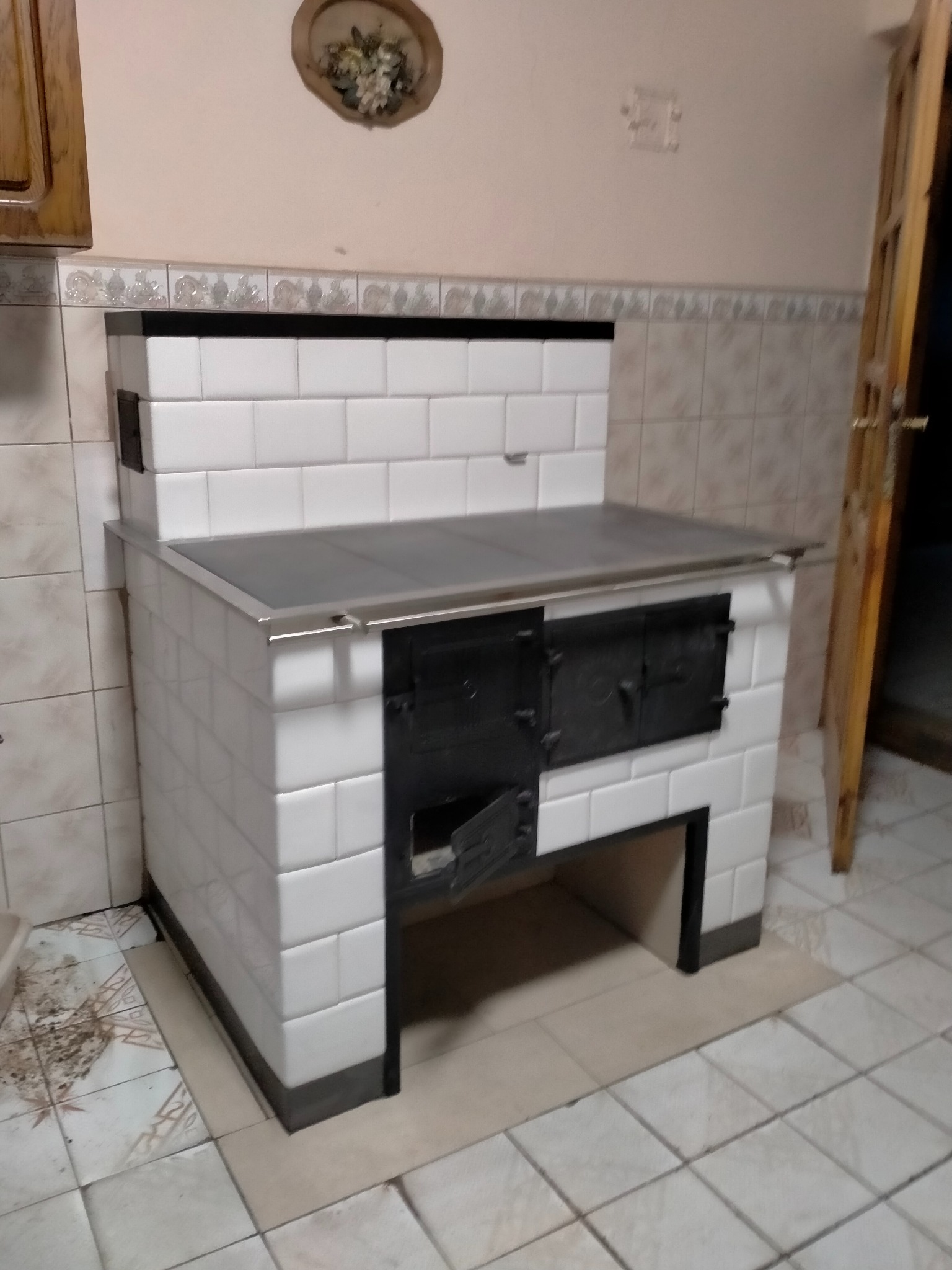 Biały piec kaflowy z otwartymi drzwiczkami paleniska, widoczne płytki na ścianie i podłodze, dekoracja kwiatowa na ścianie.