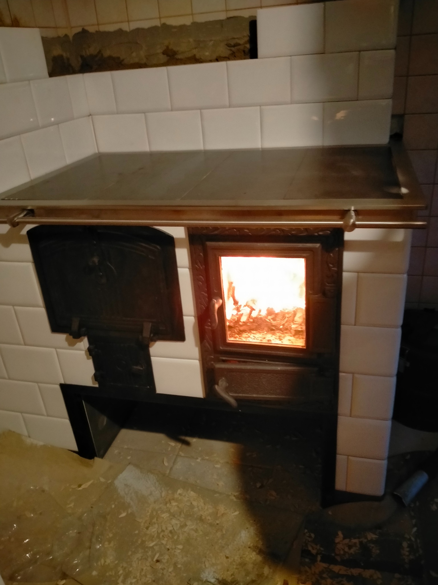 Tradycyjny piec kaflowy z otwartymi drzwiczkami, wewnątrz widoczne płomienie ognia, białe kafle na ścianach, drewniane wióry na podłodze.