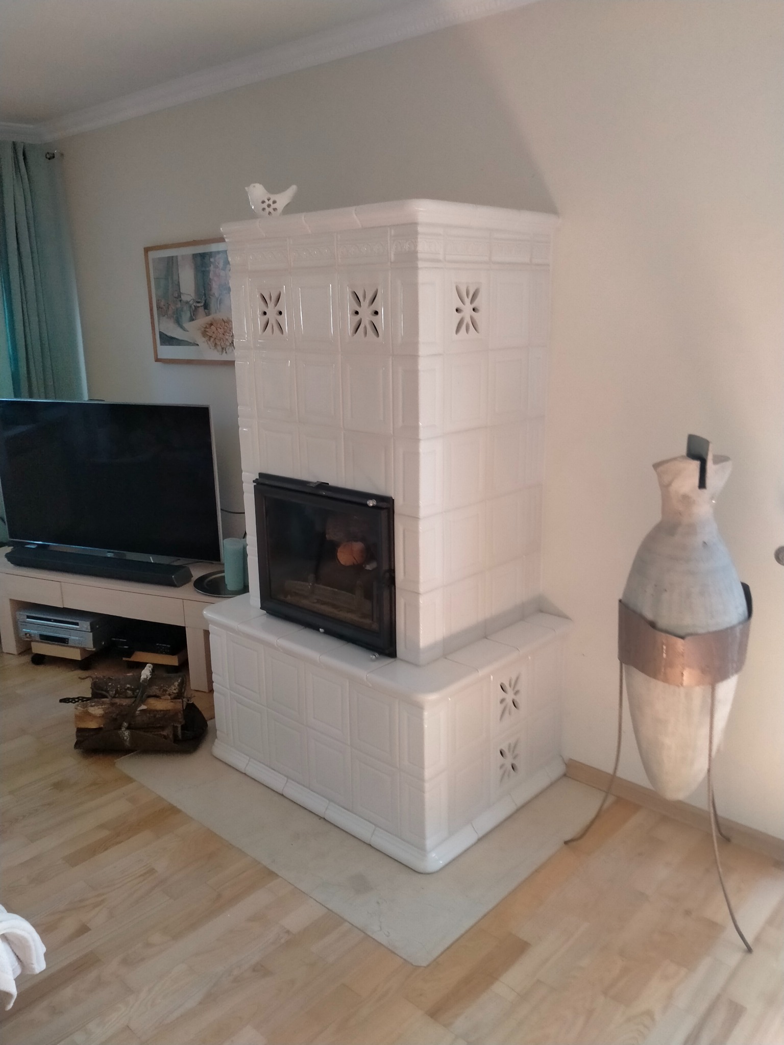 Biały, kaflowy piec z czarnym wkładem kominkowym, ozdobiony motywem kwiatowym, zwieńczony ceramiczną figurką ptaka, w salonie z drewnianą podłogą i dekoracyjną wazą