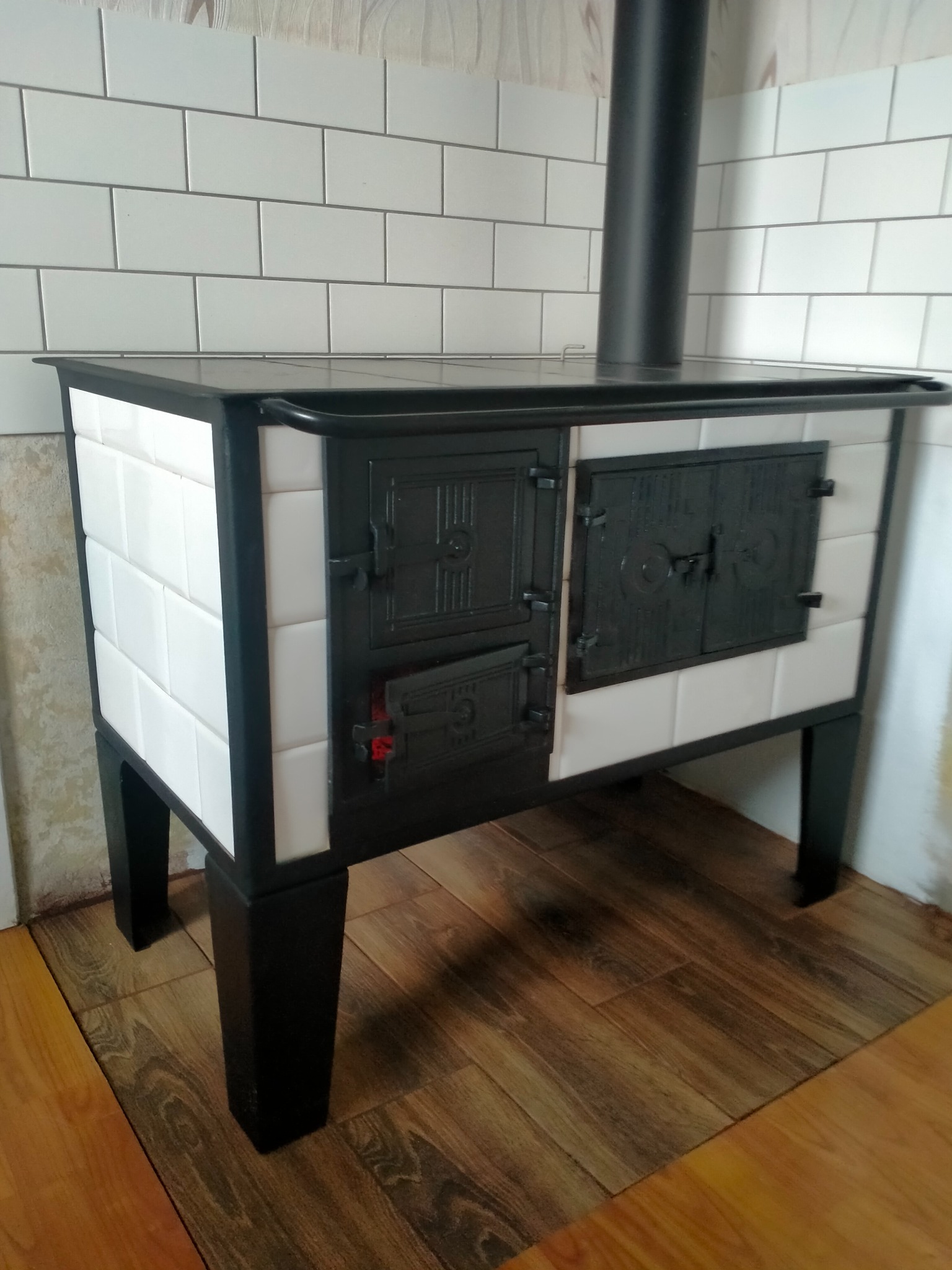 Kuchenny piec kaflowy z czarnymi drzwiczkami i rurą odprowadzającą dym, obudowany białymi kaflami, ustawiony na drewnianej podłodze.