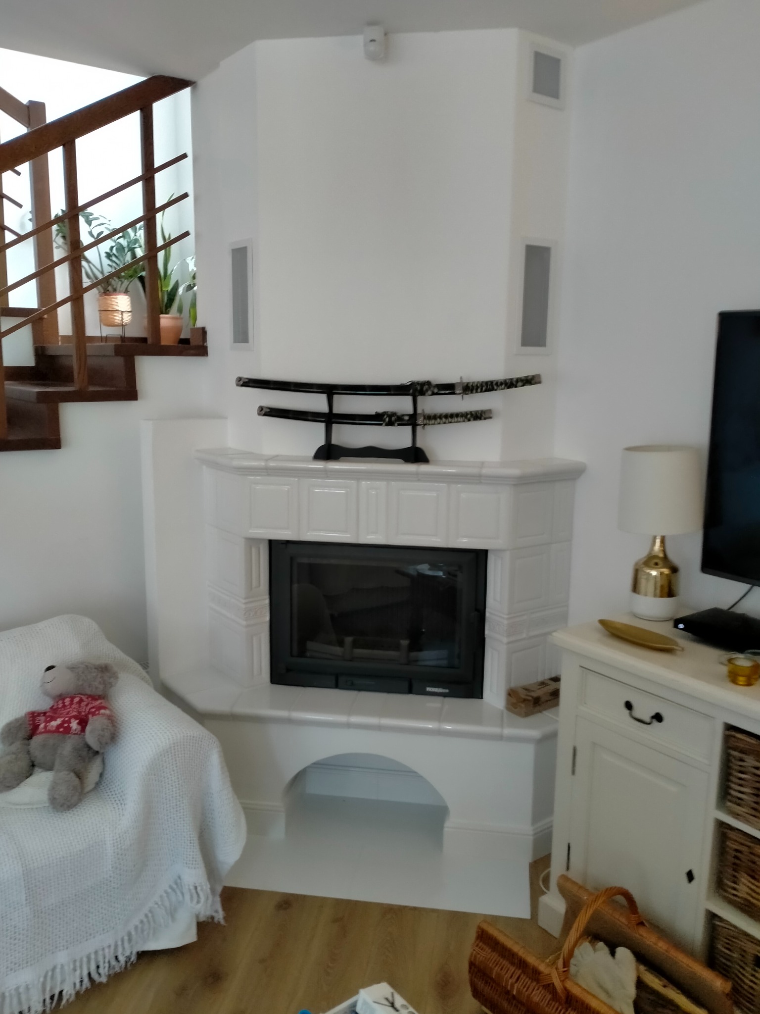 Biały kominek narożny z ceramiczną obudową, ozdobiony dekoracyjnymi katanami, umieszczony w jasnym salonie obok drewnianych schodów i białej komody z lampką.