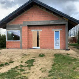 Surowy stan domu z czerwonej cegły z widocznymi otworami okiennymi i wejściowymi, betonowe nadproża, dach pokryty czarną dachówką, teren wokół w stanie naturalnym.